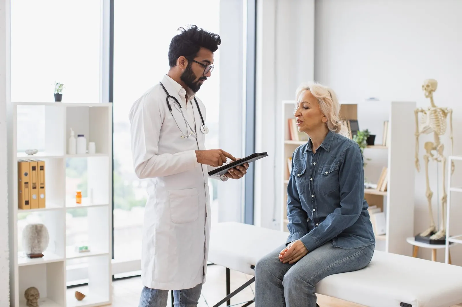 Bearded male doctor and elderly female patient sitting at exam couch.